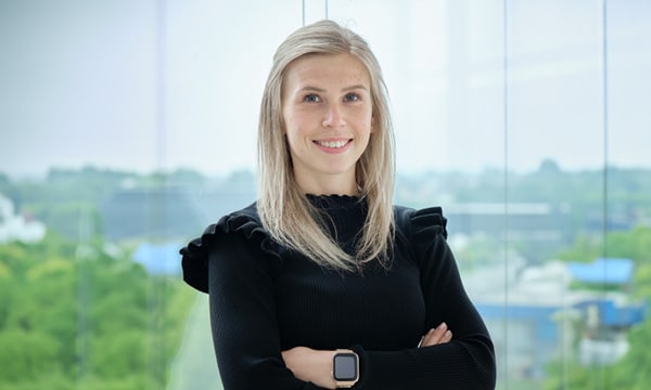 Eine Frau mit blonden Haaren, einem schwarzen Pullover und einer Smartwatch steht selbstbewusst mit verschränkten Armen da. Sie lächelt in einem Büro mit einem großen Fenster, das einen verschwommenen, grünen Außenhintergrund freigibt. Ihre Rolle beim IT-Sicherheitsdienstleister umfasst die Überwachung kritischer Aufgaben wie die Durchführung von Phishing-Tests.