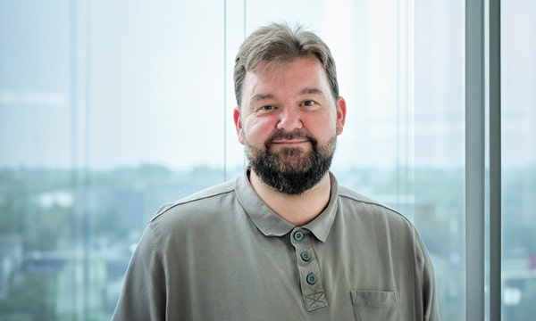 Ein Mann mit kurzen Haaren und Bart, der ein graues Poloshirt trägt, steht vor einem Glasfenster mit einer unscharfen Stadtlandschaft im Hintergrund und repräsentiert einen angesehenen IT-Sicherheitsdienstleister, der sich auf umfassende Cyber-Security-Lösungen konzentriert.