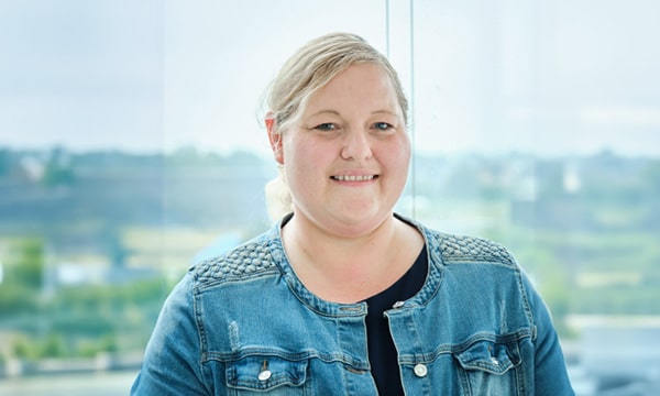 Eine Person mit hellem, zu einem Pferdeschwanz gebundenem Haar lächelt in die Kamera. Sie trägt eine blaue Jeansjacke und steht vor einem großen Fenster, im Hintergrund ist eine verschwommene Landschaft zu sehen, und denkt vielleicht über ihren nächsten Pentest für einen IT-Sicherheitsdienstleister nach.