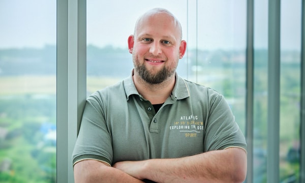 Ein Mann mit Glatze und Bart, der ein graues Poloshirt mit Text trägt, steht mit verschränkten Armen vor großen Fenstern. Die Fenster zeigen eine verschwommene Außenszene mit Grünflächen und Gebäuden im Hintergrund. Er lächelt in die Kamera und scheint ein Experte eines IT-Sicherheitsdienstleisters zu sein.