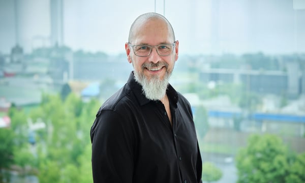 Ein Mann mit Glatze, weißem Bart und Brille lächelt und trägt ein schwarzes Hemd. Er steht vor einem großen Fenster mit einer verschwommenen Stadtlandschaft aus Bäumen und Gebäuden im Hintergrund und verkörpert die Essenz der IT-Sicherheit.