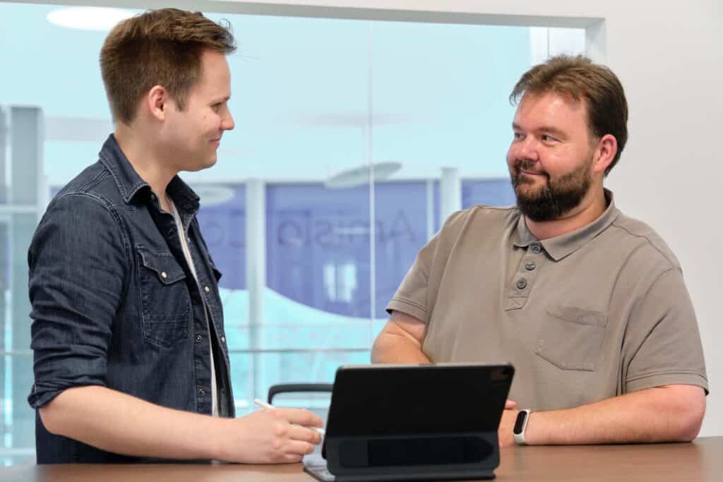 Zwei Männer unterhalten sich an einem Tisch. Einer steht und hält einen Stift und ein Notizbuch in der Hand. Er trägt ein schwarzes Hemd über einem weißen T-Shirt. Der andere Mann sitzt da, trägt ein graues Poloshirt und hat die Arme vor der Brust verschränkt. Er blickt den stehenden Mann an. Ein Laptop auf dem Tisch lässt vermuten, dass sie über Cybersicherheit sprechen oder einen Pentest planen.