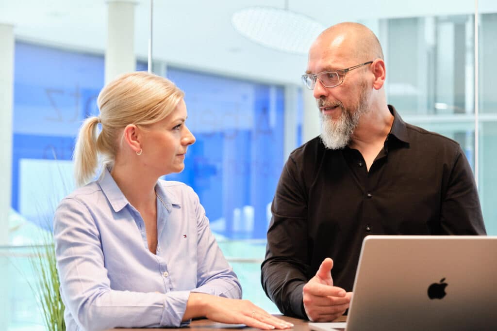 Ein Mann mit Bart und Brille, der ein schwarzes Hemd trägt, gestikuliert, während er mit einer Frau spricht, die blondes Haar zu einem Pferdeschwanz gebunden hat und ein hellblaues Hemd mit Knöpfen trägt. Sie stehen neben einem Tisch mit einem silbernen Laptop und diskutieren Cybersicherheitsstrategien in ihrer modernen, hellen Büroumgebung.