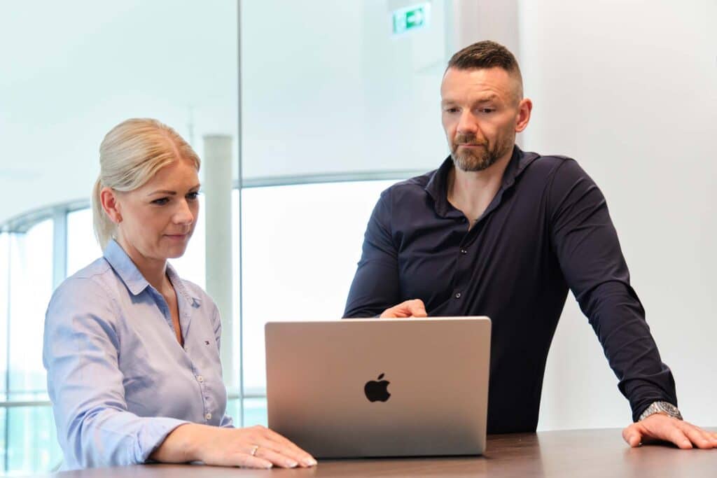 Zwei Personen, ein Mann und eine Frau, stehen an einem Tisch und schauen auf einen Laptop. Der Mann zeigt auf den Bildschirm, während die Frau zusieht. Beide sind leger gekleidet. Sie scheinen sich in einer modernen Büroumgebung zu befinden und führen möglicherweise eine IT-Sicherheitsberatung durch oder überprüfen die Ergebnisse eines Phishing-Tests.