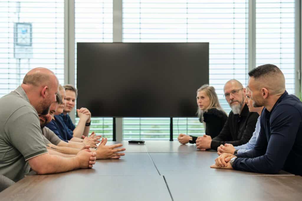 Acht Personen, Männer und Frauen, sitzen an einem Konferenztisch und diskutieren über Cybersicherheit. Im Hintergrund steht ein großer Monitor vor einem Fenster, dessen Jalousien teilweise geschlossen sind und so natürliches, diffuses Licht hereinlassen. Die Stimmung wirkt konzentriert und kooperativ.