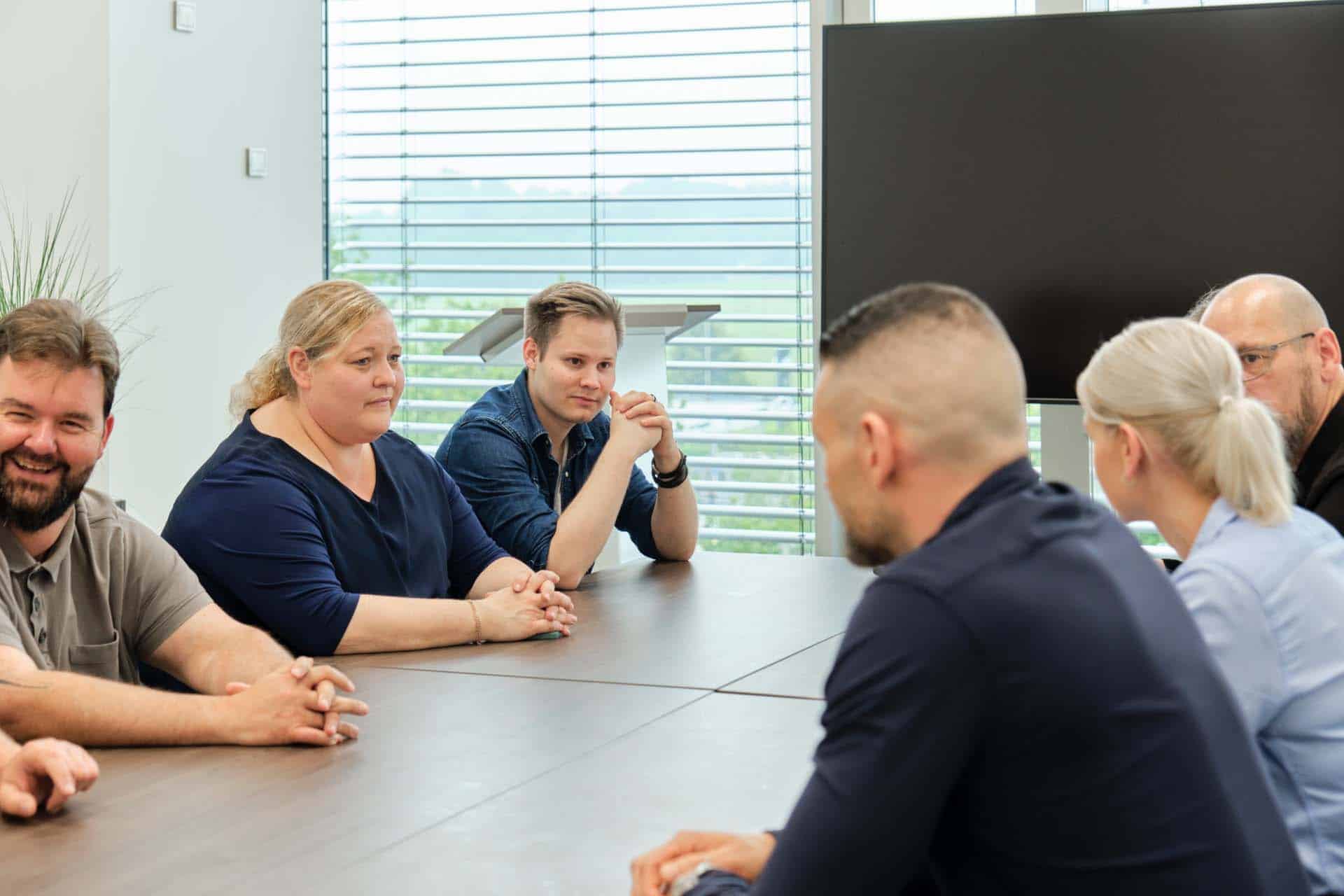 Eine Gruppe von sechs Personen sitzt um einen Konferenztisch und berät über IT-Sicherheit. Eine Frau steht in der Mitte, die anderen am Tisch blicken aufmerksam. Im Hintergrund sind ein großes Fenster mit Jalousien und ein großer Fernsehbildschirm zu sehen.