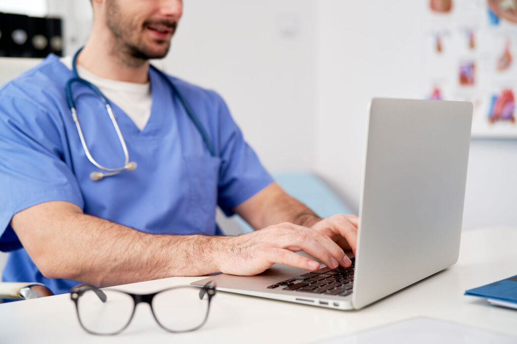 Ein Arzt im blauen Kittel und mit einem Stethoskop um den Hals sitzt an einem Schreibtisch und tippt auf einem Laptop. Im Vordergrund liegt eine Brille auf dem Schreibtisch, im Hintergrund sind Krankenakten zu sehen.