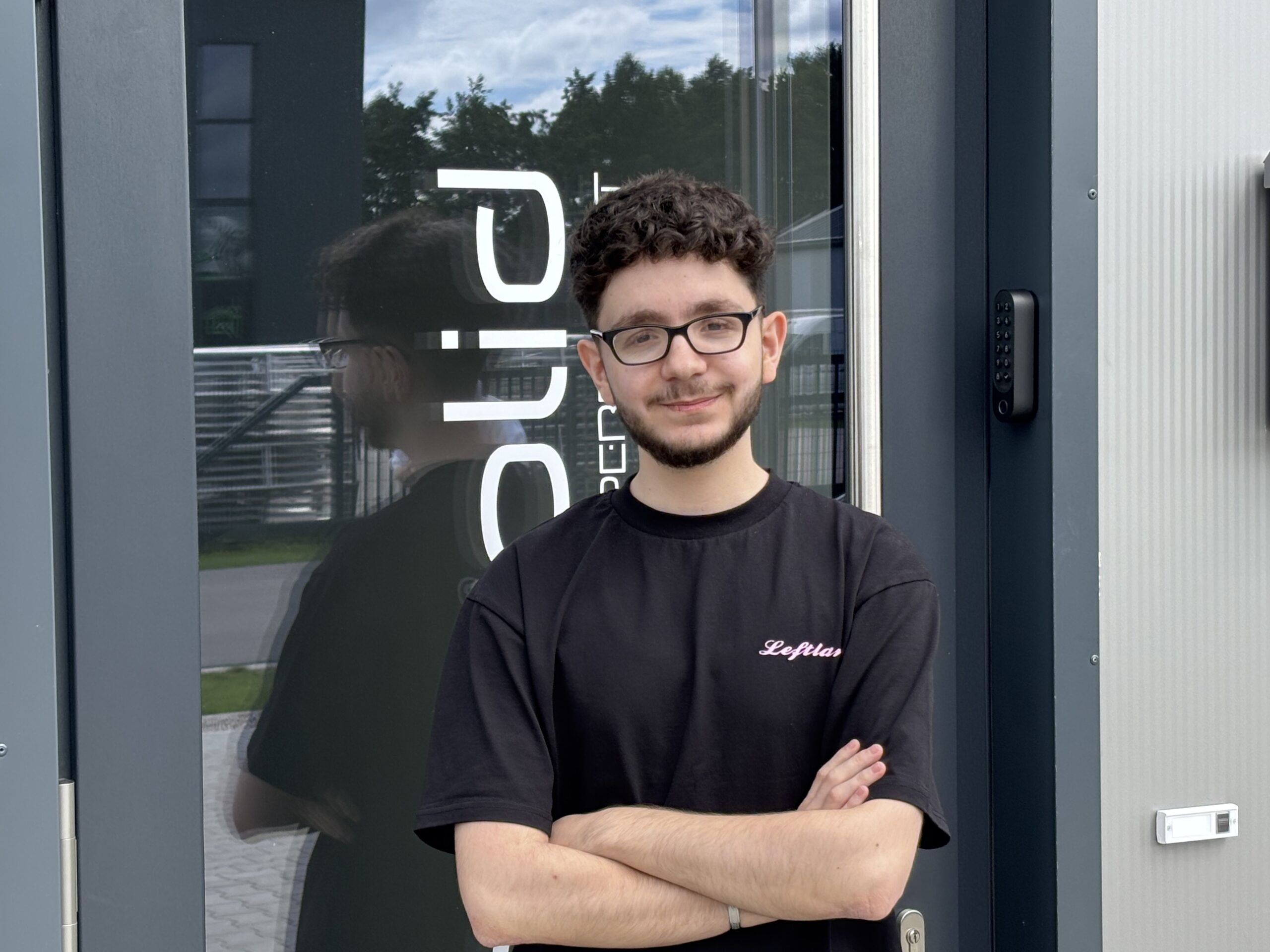 Ein junger Mann mit kurzen dunklen Haaren, Brille und Bart steht vor einem modernen Gebäude, verschränkt die Arme und lächelt in die Kamera. Er trägt ein schwarzes T-Shirt mit der Aufschrift "Leflair".