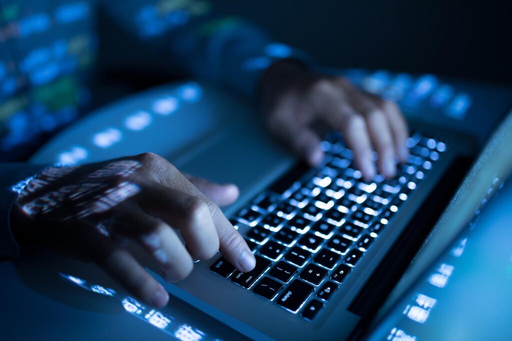 Nahaufnahme von Händen, die in einer dunklen Umgebung auf einer Laptoptastatur tippen, wobei blaues Licht und codeähnliche Reflektionen auf die Hände und die Tastatur projiziert werden.